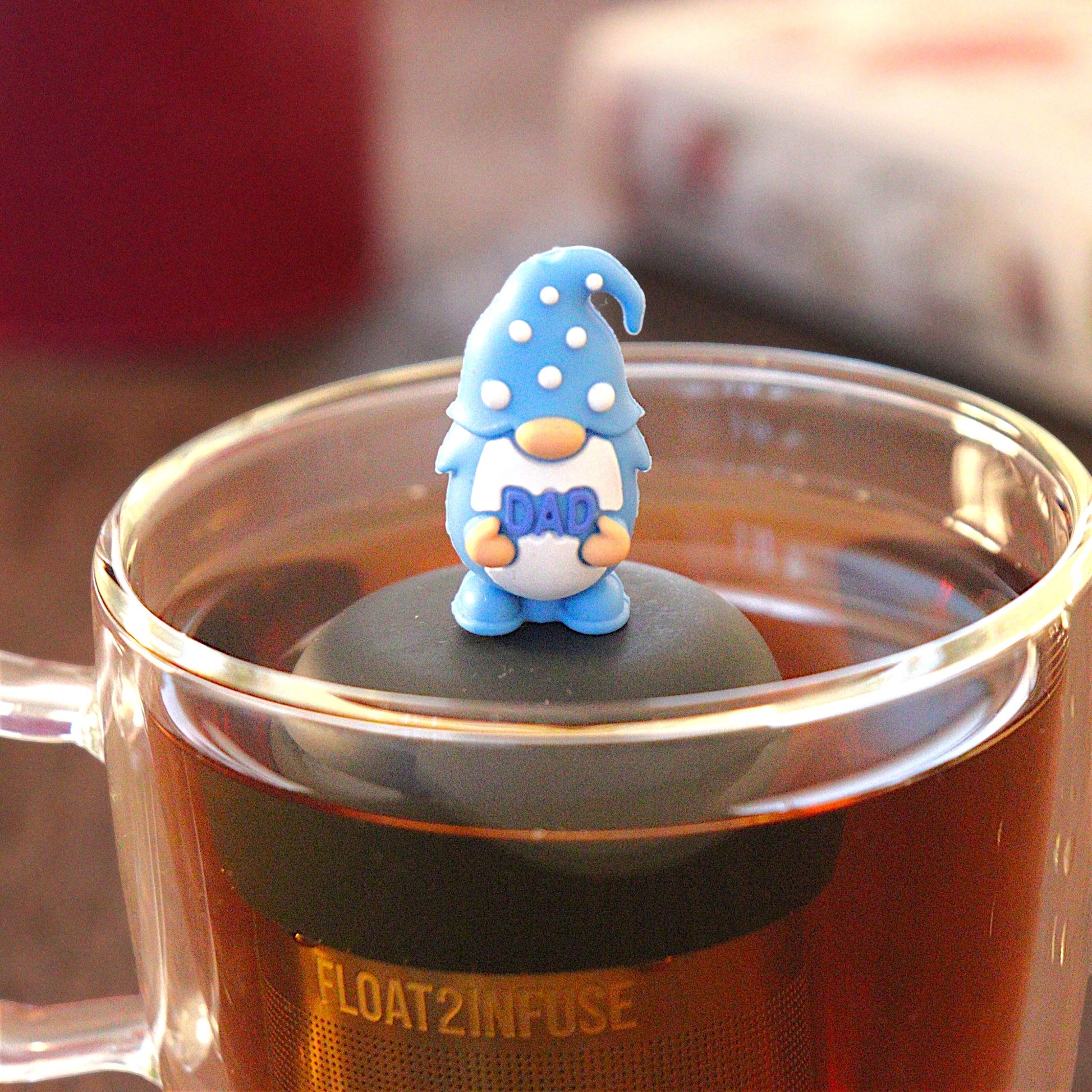 blue gnome that holding the word "dad" tea infuser for loose leaf tea  with a fine mesh stainless steel strainer that reads FLOAT2INFUSE on a black base floating in a glass tea mug with tea kettle in the background