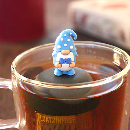 blue gnome that holding the word "dad" tea infuser for loose leaf tea  with a fine mesh stainless steel strainer that reads FLOAT2INFUSE on a black base floating in a glass tea mug with tea kettle in the background
