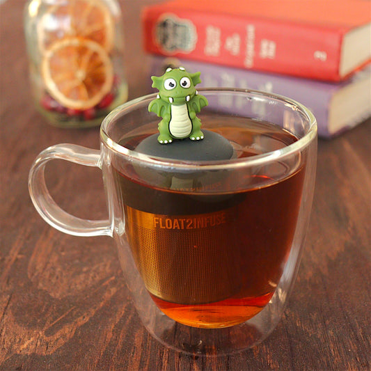 dark green dragon tea infuser for loose leaf tea  with a fine mesh stainless steel tea strainer that reads FLOAT2INFUSE on a black base floating in a glass tea mug with loose leaf tea and books in the background