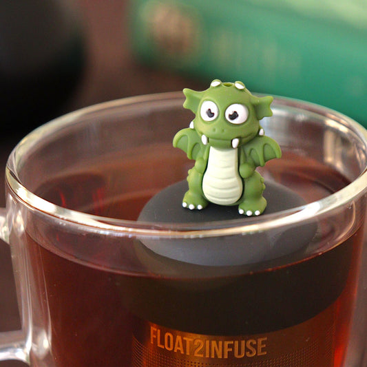 dark green dragon tea infuser for loose leaf tea  with a fine mesh stainless steel tea strainer that reads FLOAT2INFUSE on a black base floating in a glass tea mug with loose leaf tea and books in the background