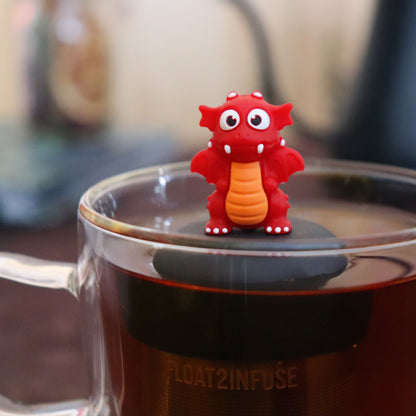 red dragon animal tea infuser for loose leaf tea  with a fine mesh stainless steel tea infuser that reads FLOAT2INFUSE on a black base floating in a glass tea mug with tea kettle in the background