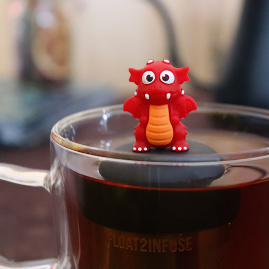 red dragon animal tea infuser for loose leaf tea  with a fine mesh stainless steel tea infuser that reads FLOAT2INFUSE on a black base floating in a glass tea mug with tea kettle in the background