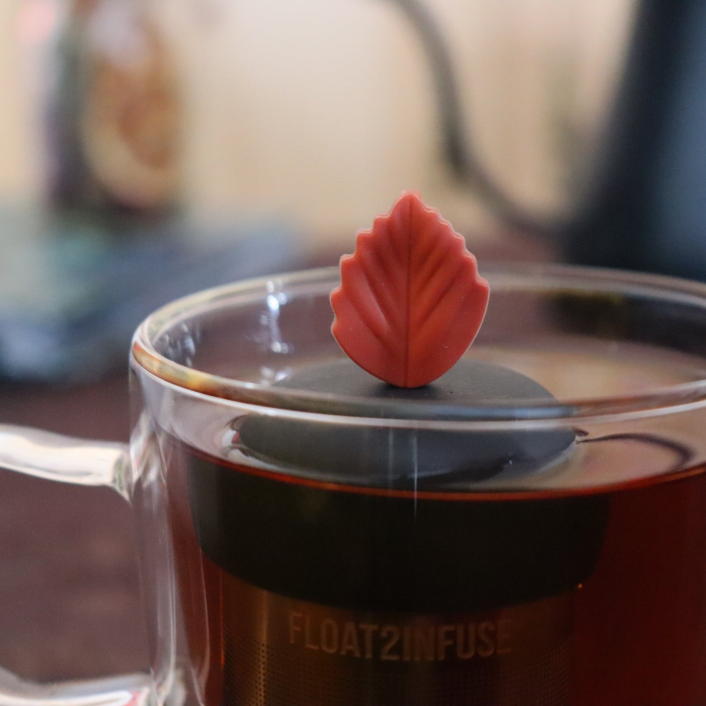 maroon fall leaf tea infuser for loose leaf tea  with a fine mesh stainless steel strainer that reads FLOAT2INFUSE on a black base floating in a glass tea mug with tea kettle in the background