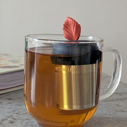maroon leaf tea infuser for loose leaf tea  with a fine mesh stainless steel tea infuser that reads FLOAT2INFUSE on a black base floating in a glass tea mug with books and loose leaf tea in the background