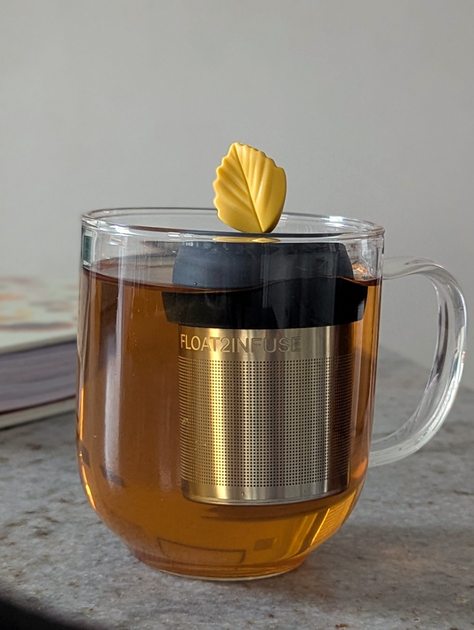 yellow fall leaf tea infuser for loose leaf tea  with a fine mesh stainless steel strainer that reads FLOAT2INFUSE on a black base floating in a glass tea mug with a book in the background