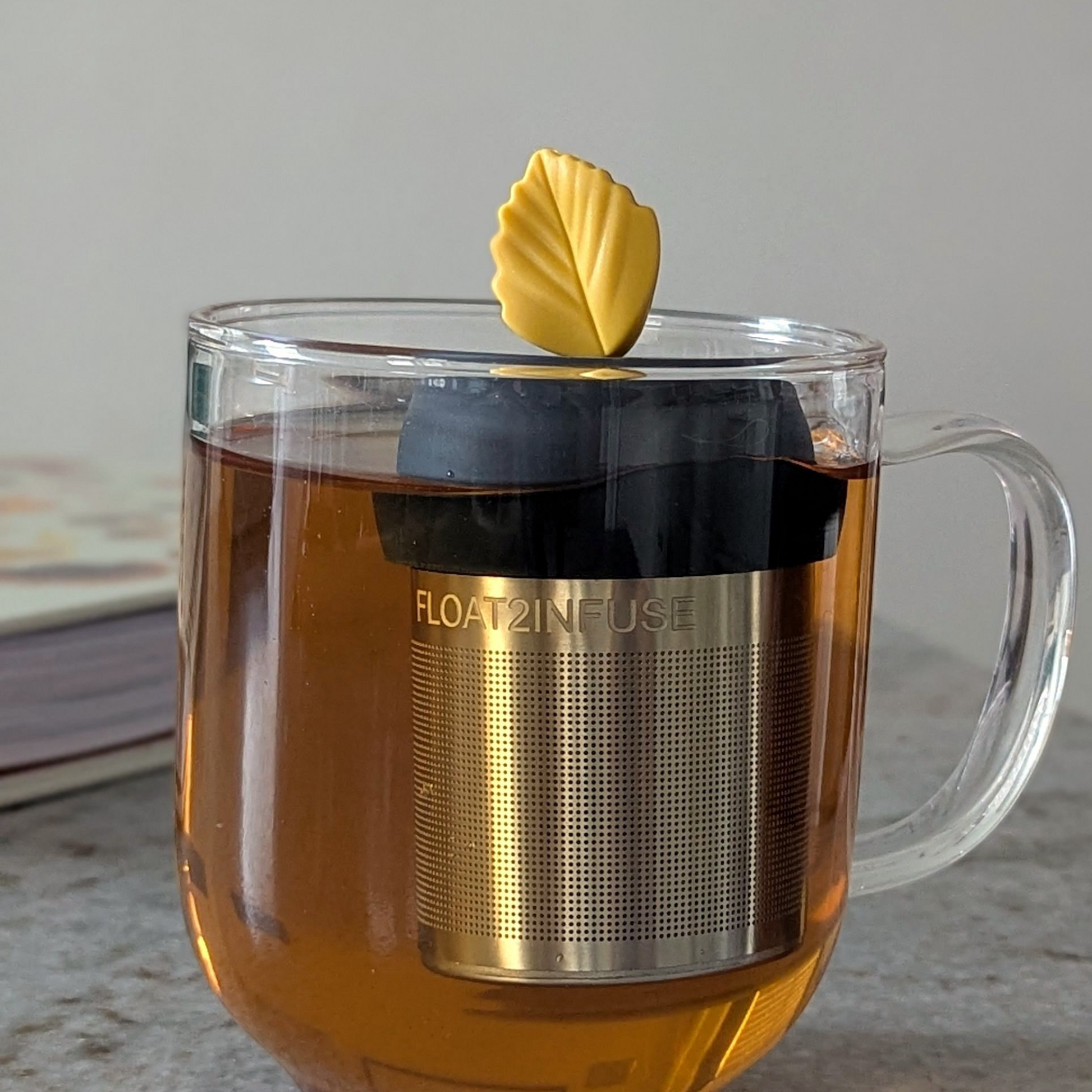 yellow fall leaf tea infuser for loose leaf tea  with a fine mesh stainless steel strainer that reads FLOAT2INFUSE on a black base floating in a glass tea mug with a book in the background