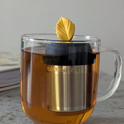 yellow fall leaf tea infuser for loose leaf tea  with a fine mesh stainless steel strainer that reads FLOAT2INFUSE on a black base floating in a glass tea mug with a book in the background