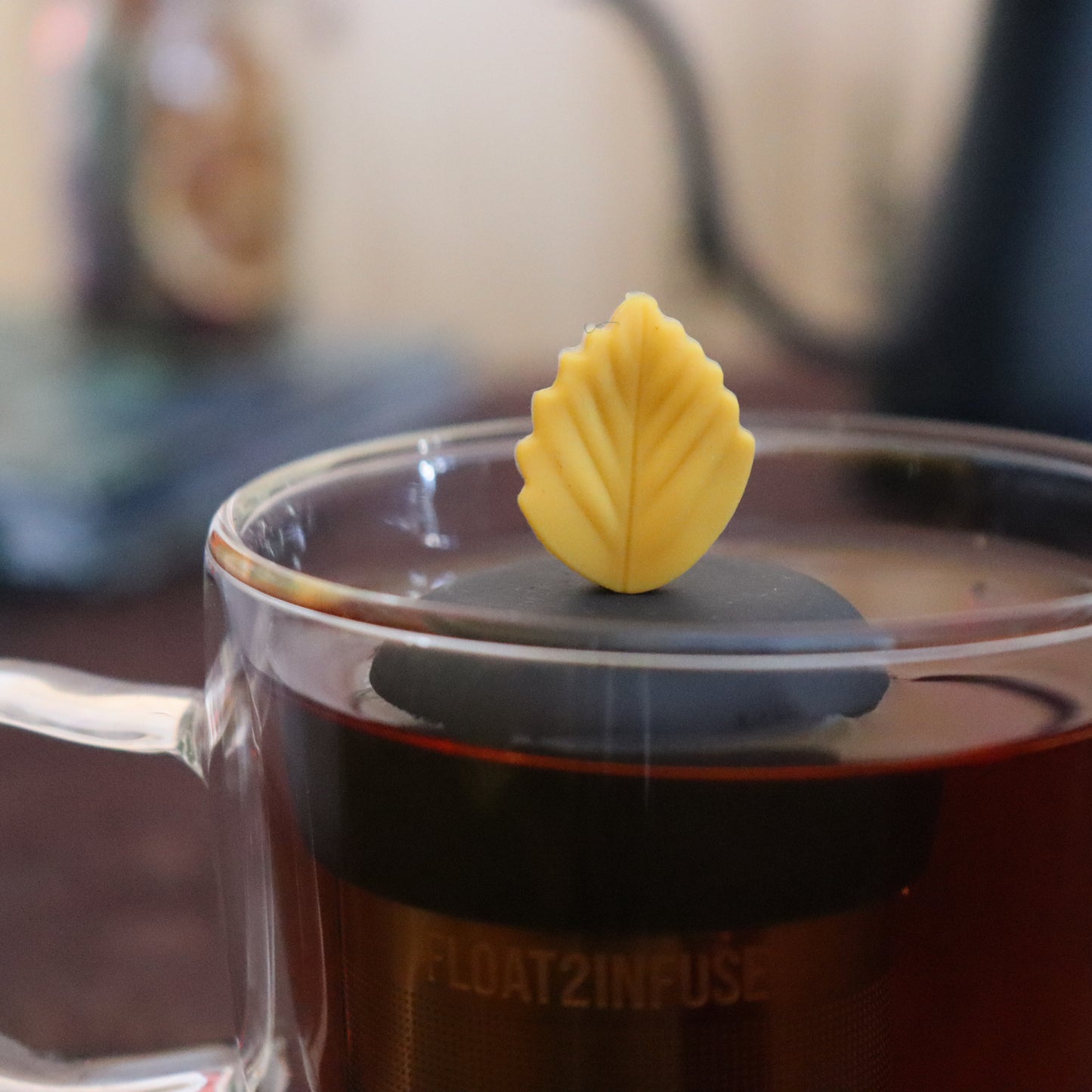 yellow fall leaf tea infuser for loose leaf tea  with a fine mesh stainless steel strainer that reads FLOAT2INFUSE on a black base floating in a glass tea mug with tea kettle in the background