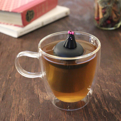 blushing black ghost with pink bow tea infuser for loose leaf tea  with a fine mesh stainless steel tea infuser that reads FLOAT2INFUSE on a black base floating in a glass tea mug with books and loose leaf tea in the background