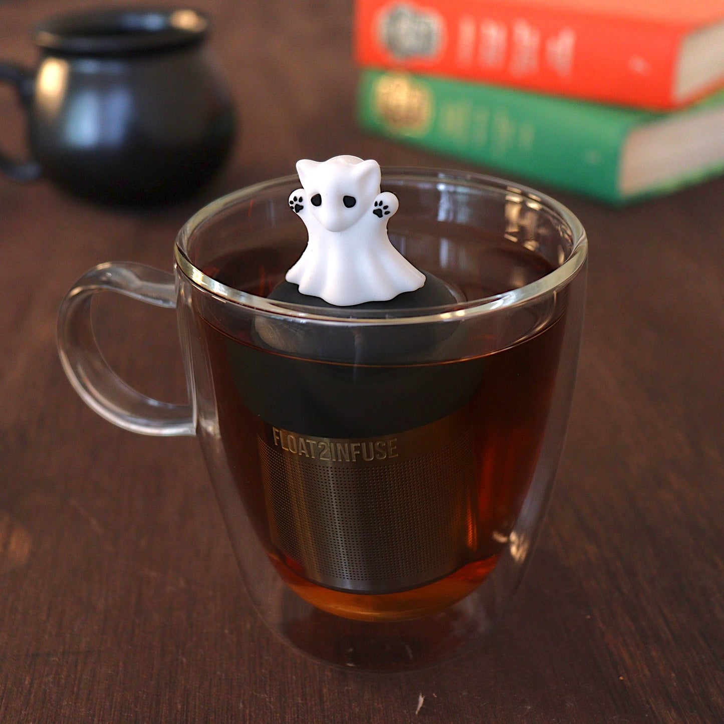 Adorable white cat ghost was big black eyes and black paws tea infuser for loose leaf tea with a fine mesh stainless steel tea strainer 
that reads FLOAT2INFUSE on a black base with a black cauldron tea mug and books in the background