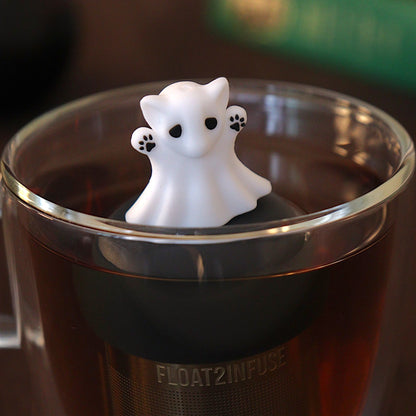 Adorable white cat ghost was big black eyes and black paws tea infuser for loose leaf tea with a fine mesh stainless steel tea strainer 
that reads FLOAT2INFUSE on a black base with a black cauldron tea mug and books in the background