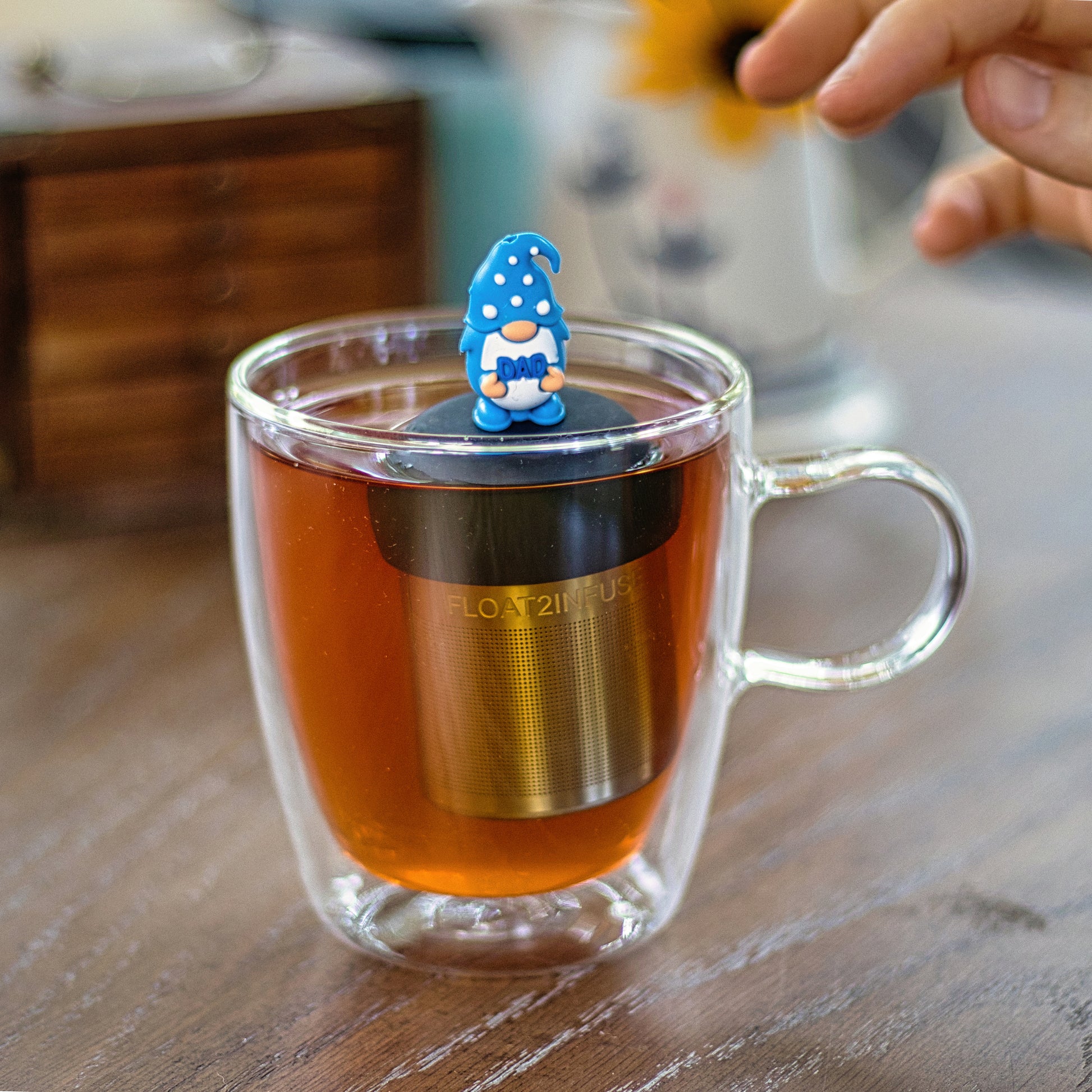 gnome with long white beard wearing blue with white polka dots holds text "dad" tea infuser for loose leaf tea with a fine mesh stainless steel tea infuser that reads FLOAT2INFUSE on a black base  floating in a glass tea mug with  drink pitcher in the background
