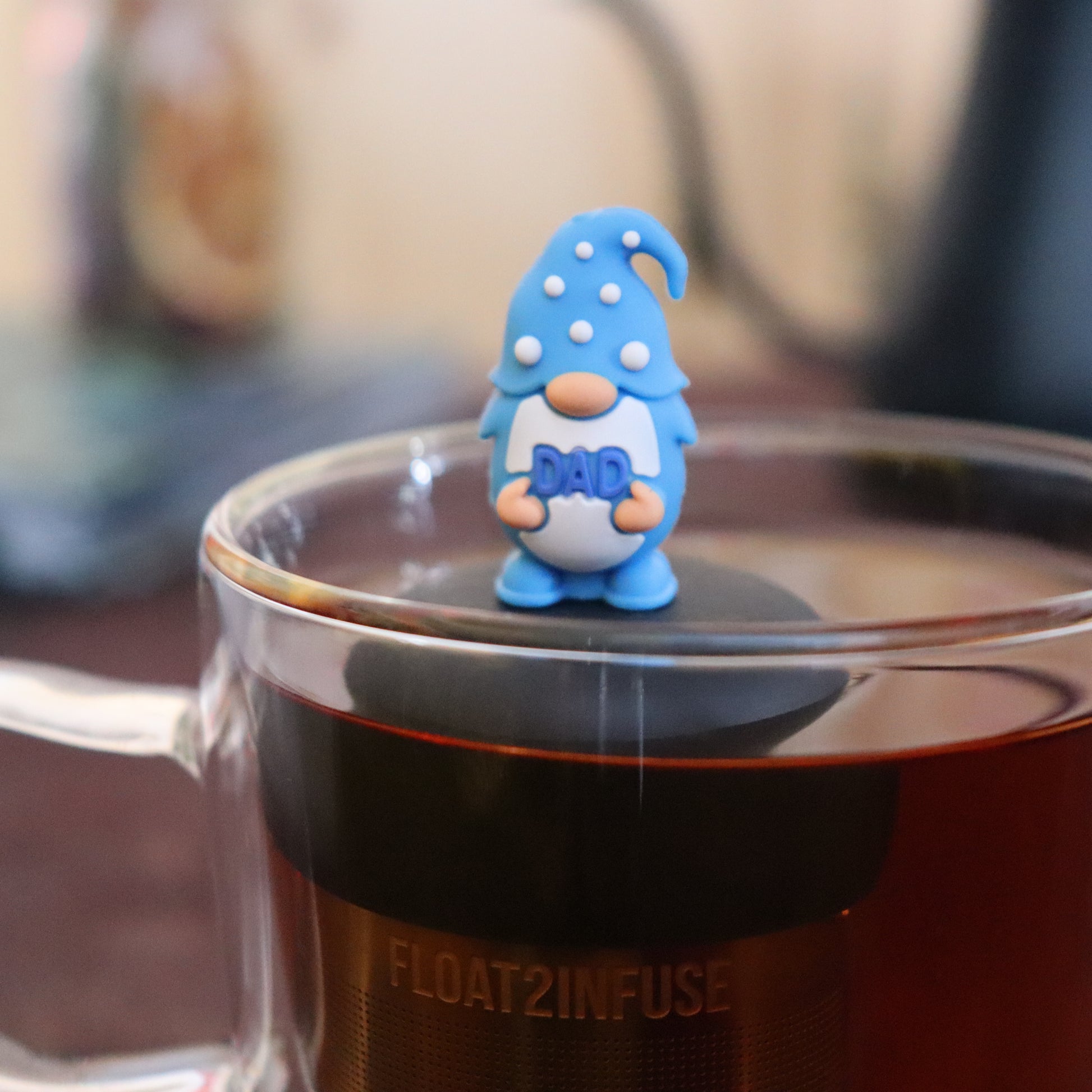 blue gnome that holding the word "dad" tea infuser for loose leaf tea  with a fine mesh stainless steel strainer that reads FLOAT2INFUSE on a black base floating in a glass tea mug with tea kettle in the background
