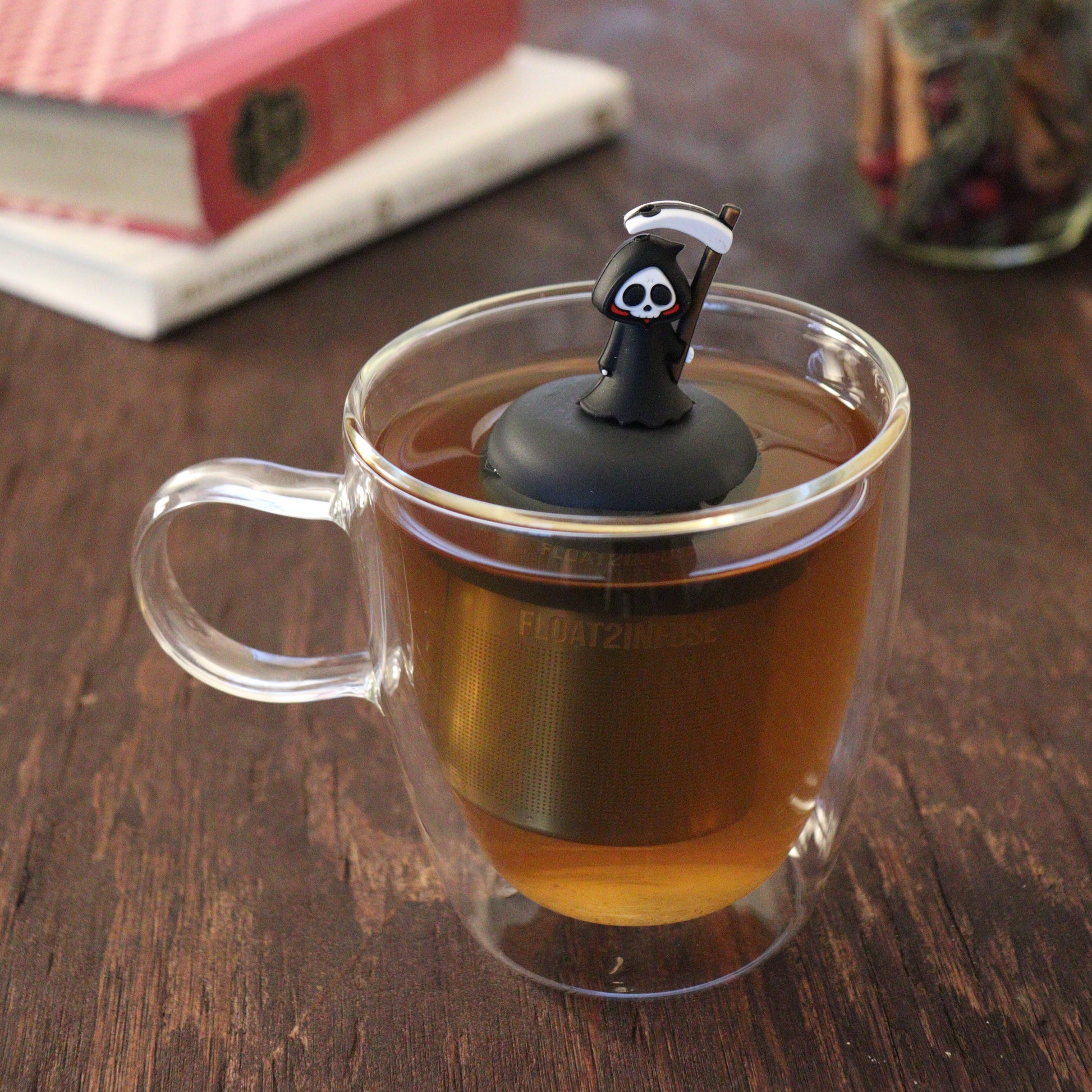 black grim reaper tea infuser for loose leaf tea with a fine mesh stainless steel tea infuser that reads FLOAT2INFUSE on a black base floating in a glass tea mug with books and loose leaf tea in the background
