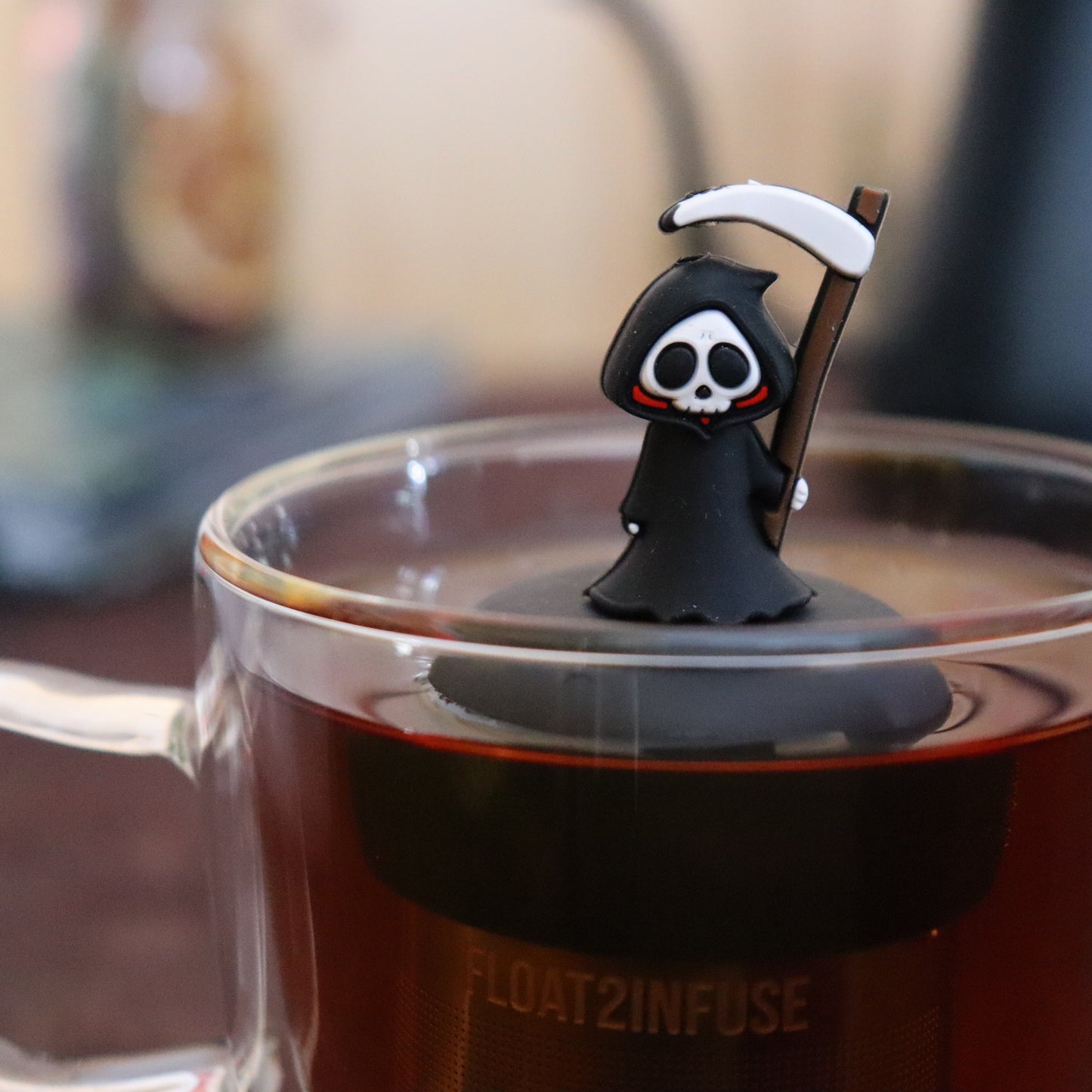 black grim reaper tea infuser for loose leaf tea  with a fine mesh stainless steel strainer that reads FLOAT2INFUSE on a black base floating in a glass tea mug with tea kettle in the background
