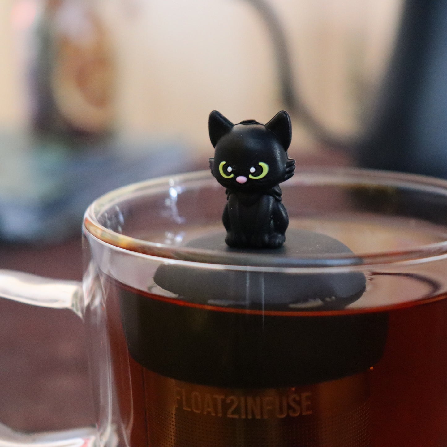 black cat animal tea infuser for loose leaf tea  with a fine mesh stainless steel tea infuser that reads FLOAT2INFUSE on a black base floating in a glass tea mug with tea kettle in the background