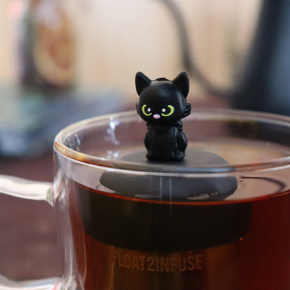 black cat animal tea infuser for loose leaf tea  with a fine mesh stainless steel tea infuser that reads FLOAT2INFUSE on a black base floating in a glass tea mug with tea kettle in the background