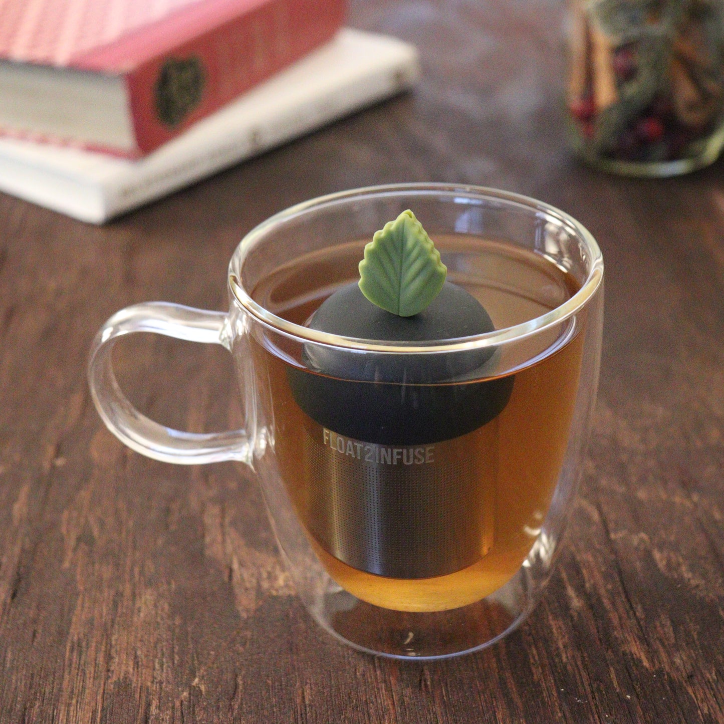 green herb leaf tea infuser for loose leaf tea on a black base with a fine mesh stainless steel tea infuser that reads FLOAT2INFUSE floating in a glass tea mug with books and loose leaf tea in the background