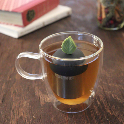 green herb leaf tea infuser for loose leaf tea on a black base with a fine mesh stainless steel tea infuser that reads FLOAT2INFUSE floating in a glass tea mug with books and loose leaf tea in the background