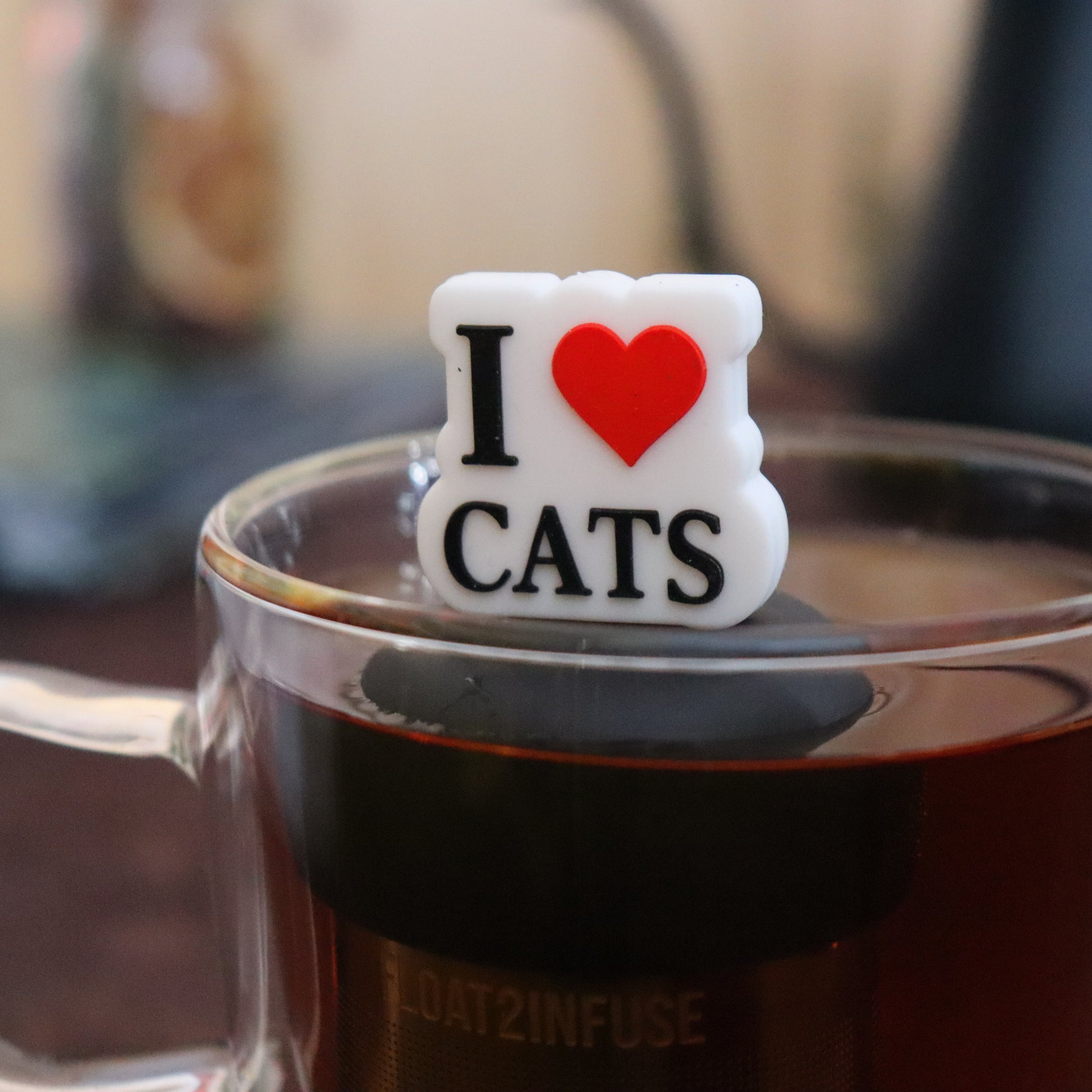 black text with white boarder and big red heart that reads "I love cats" tea infuser for loose leaf tea on a black base with a fine mesh stainless steel strainer that reads FLOAT2INFUSE floating in a glass tea mug with tea kettle in the background
