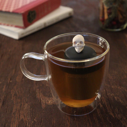 ivory skull tea infuser for loose leaf tea with a fine mesh stainless steel tea infuser that reads FLOAT2INFUSE  on a black base floating in a glass tea mug with books and loose leaf tea in the background