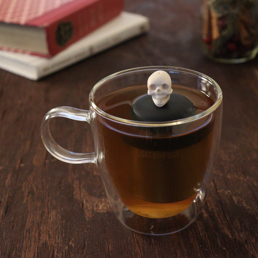 ivory skull tea infuser for loose leaf tea with a fine mesh stainless steel tea infuser that reads FLOAT2INFUSE  on a black base floating in a glass tea mug with books and loose leaf tea in the background