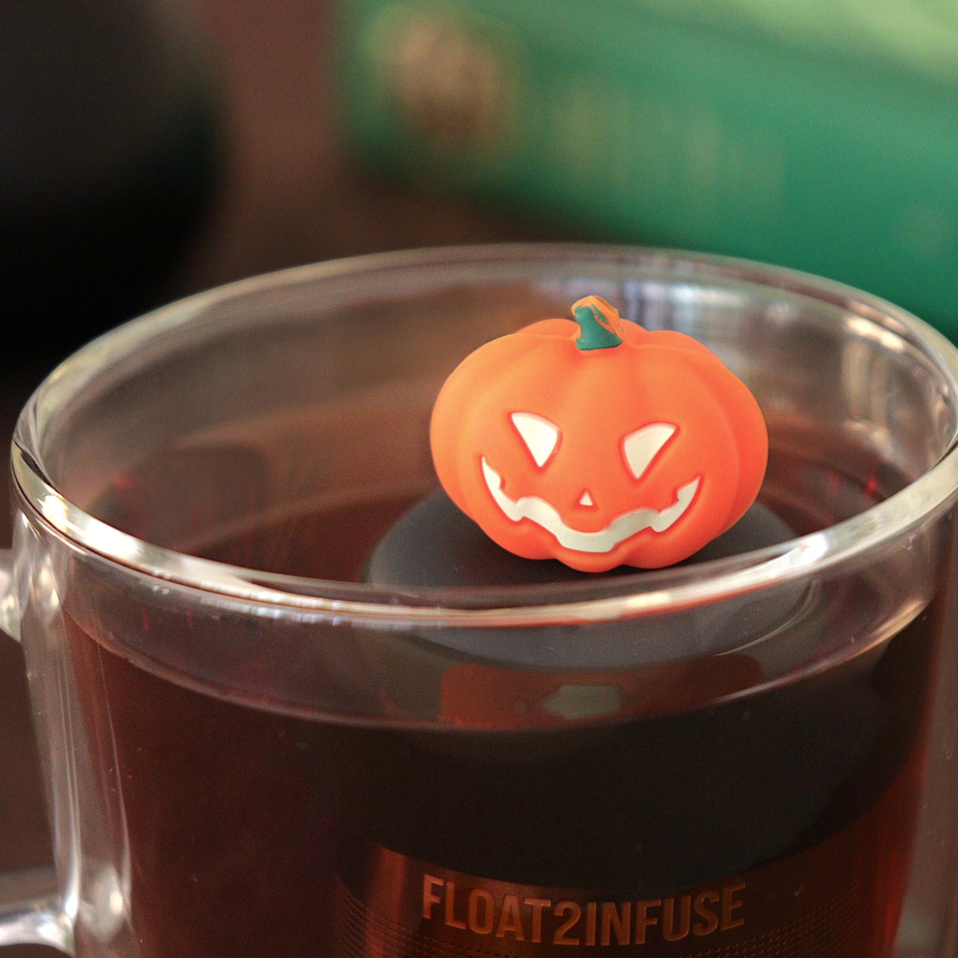 orange jack-o-lantern tea infuser for loose leaf tea with a fine mesh stainless steel tea strainer that reads FLOAT2INFUSE on a black base   floating in a glass tea mug with a cauldron and books in the background
