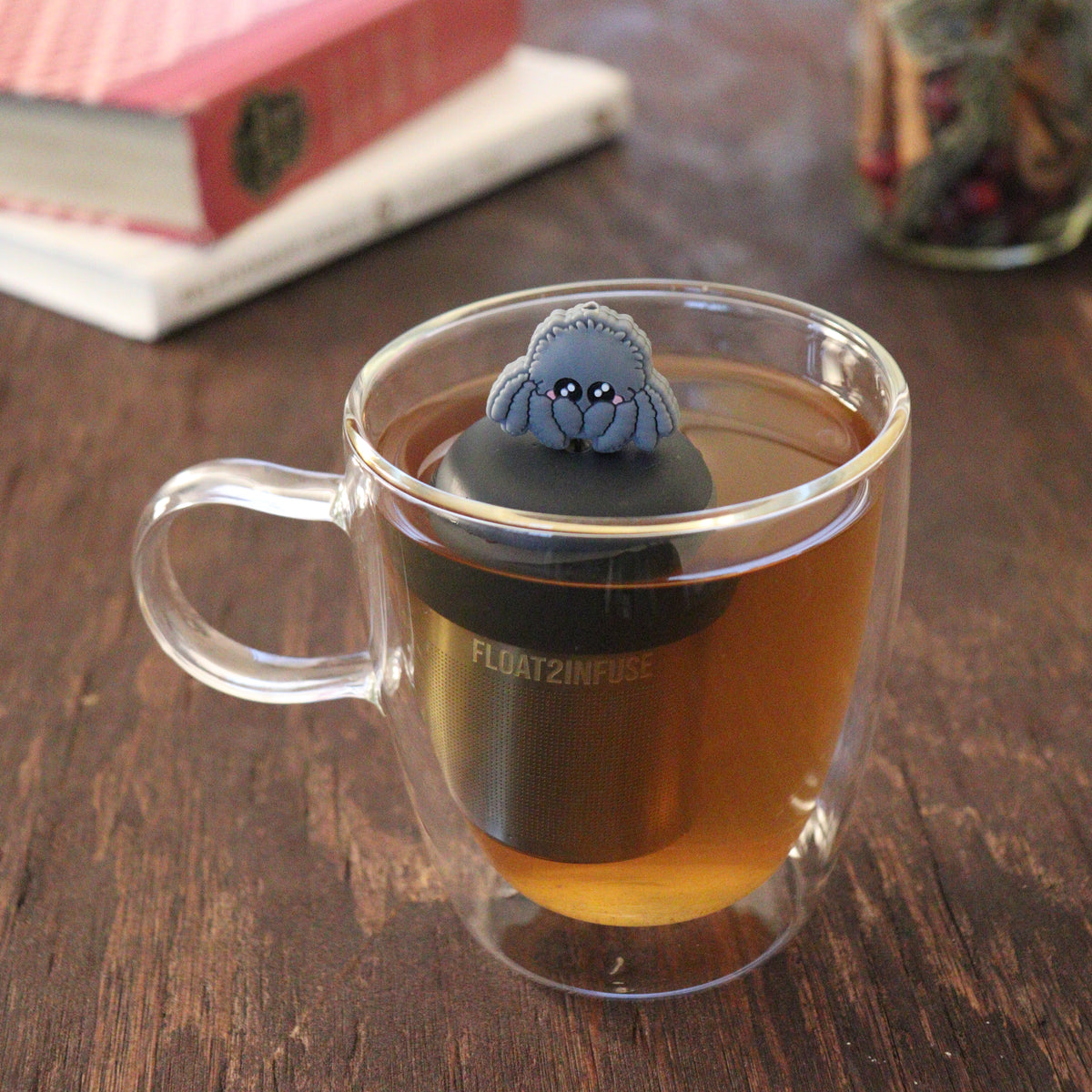 blushing grey jumping spider animal tea infuser for loose leaf tea  with a fine mesh stainless steel tea infuser that reads FLOAT2INFUSE on a black base floating in a glass tea mug with books and loose leaf tea in the background
