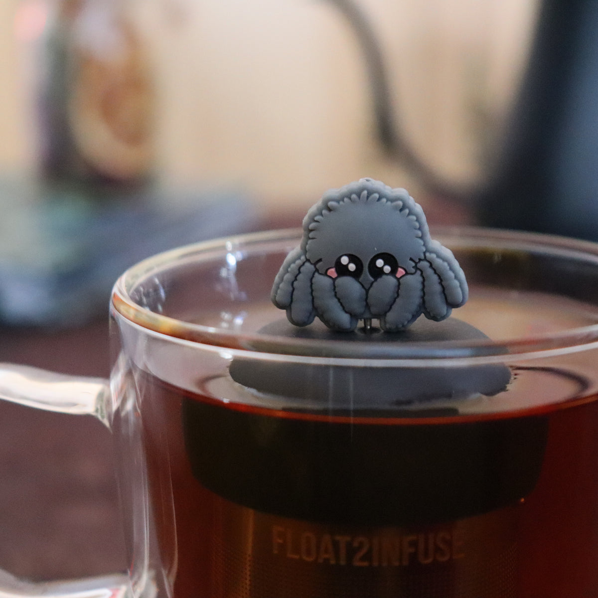 blushing grey jumping spider animal tea infuser for loose leaf tea  with a fine mesh stainless steel tea infuser that reads FLOAT2INFUSE on a black base floating in a glass tea mug with tea kettle in the background