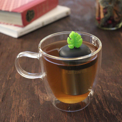 lime green monstera leaf tea infuser for loose leaf tea  with a fine mesh stainless steel tea infuser that reads FLOAT2INFUSE on a black base floating in a glass tea mug with books and loose leaf tea in the background