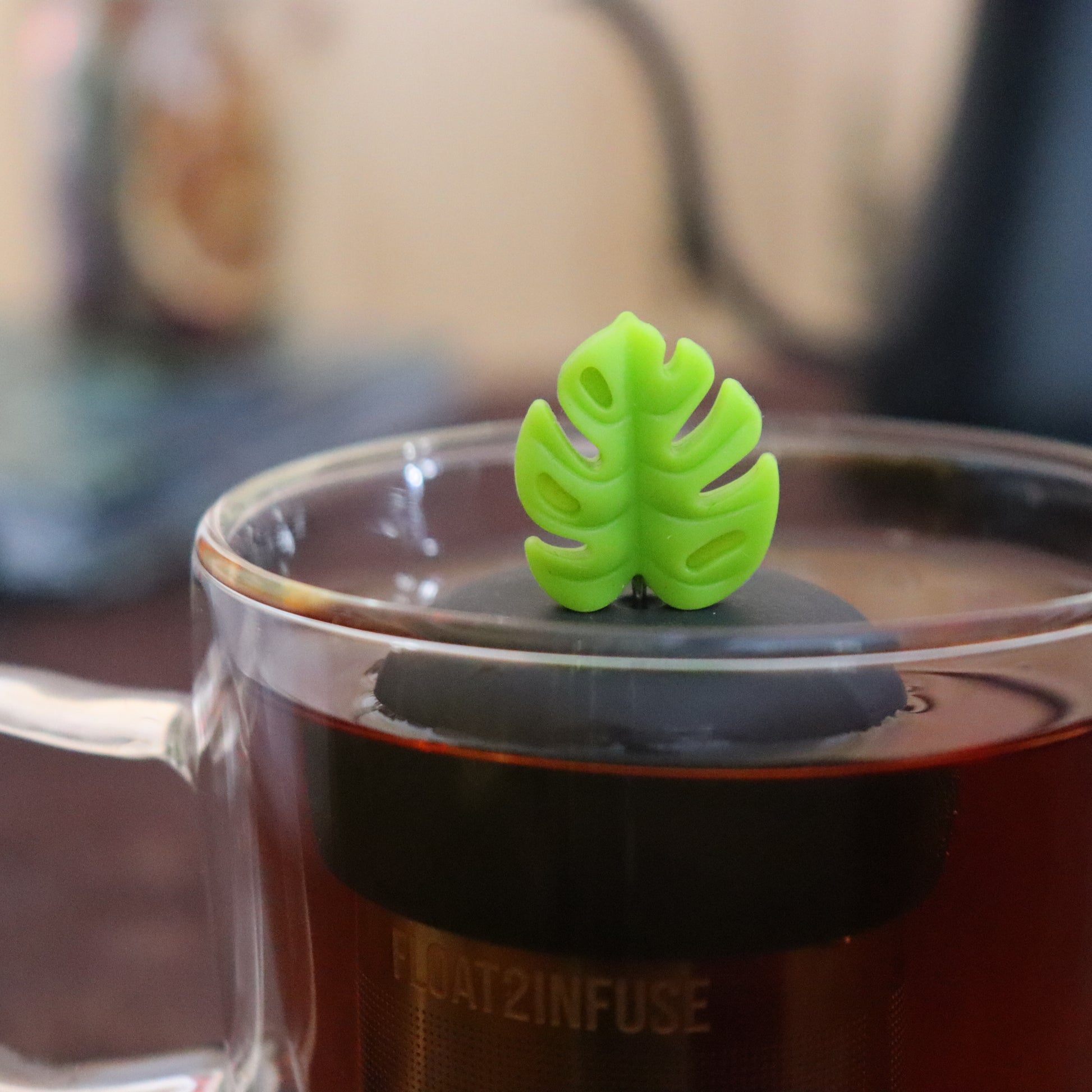 lime green monstera leaf tea infuser for loose leaf tea  with a fine mesh stainless steel strainer that reads FLOAT2INFUSE on a black base floating in a glass tea mug with tea kettle in the background