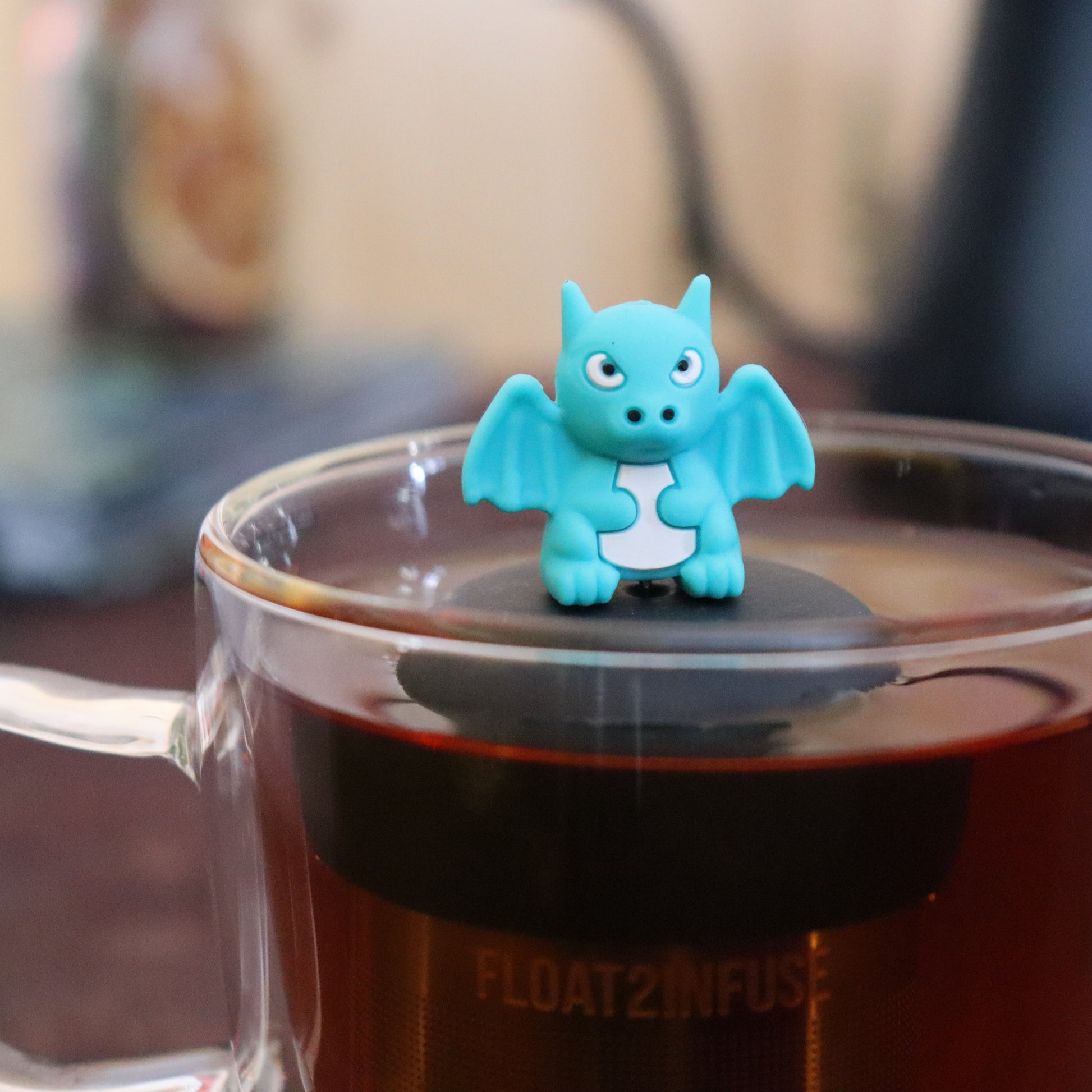 teal dragon animal tea infuser for loose leaf tea  with a fine mesh stainless steel tea infuser that reads FLOAT2INFUSE on a black base floating in a glass tea mug with tea kettle in the background