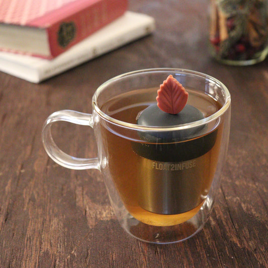 maroon leaf tea infuser for loose leaf tea  with a fine mesh stainless steel tea infuser that reads FLOAT2INFUSE on a black base floating in a glass tea mug with books and loose leaf tea in the background