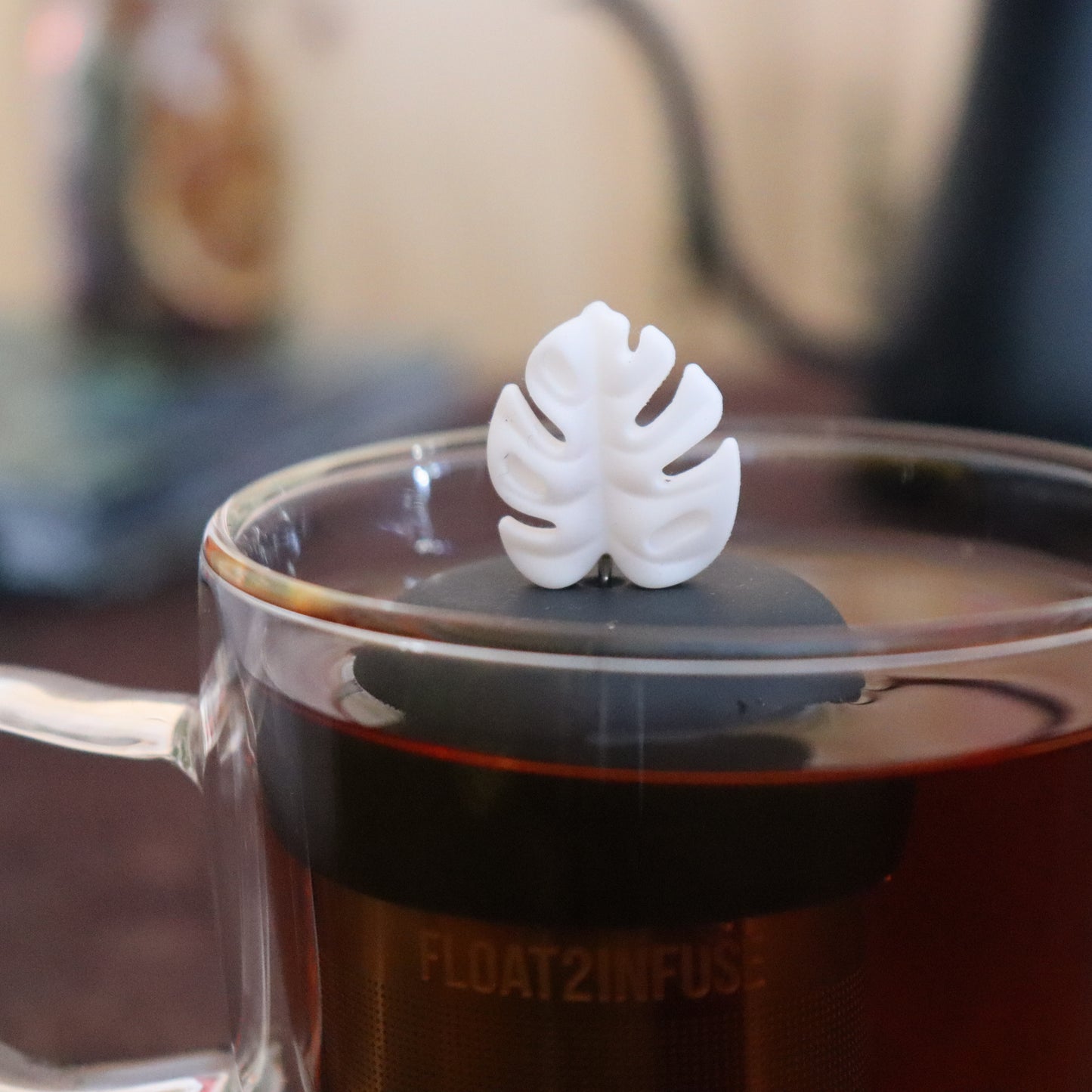 white monstera leaf tea infuser for loose leaf tea on a black base with a fine mesh stainless steel strainer that reads FLOAT2INFUSE floating in a glass tea mug with tea kettle in the background