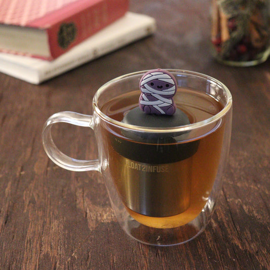 purple mummy tea infuser for loose leaf tea  with a fine mesh stainless steel tea infuser that reads FLOAT2INFUSE on a black base floating in a glass tea mug with books and loose leaf tea in the background