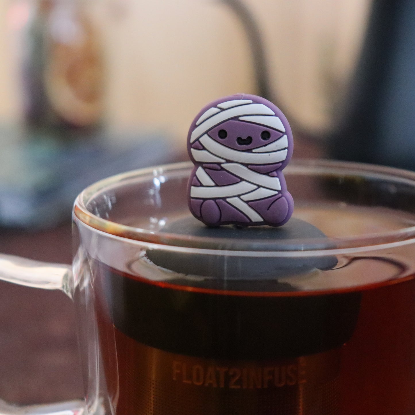 purple mummy animal tea infuser for loose leaf tea with a fine mesh stainless steel tea infuser that reads FLOAT2INFUSE on a black base  floating in a glass tea mug with tea kettle in the background