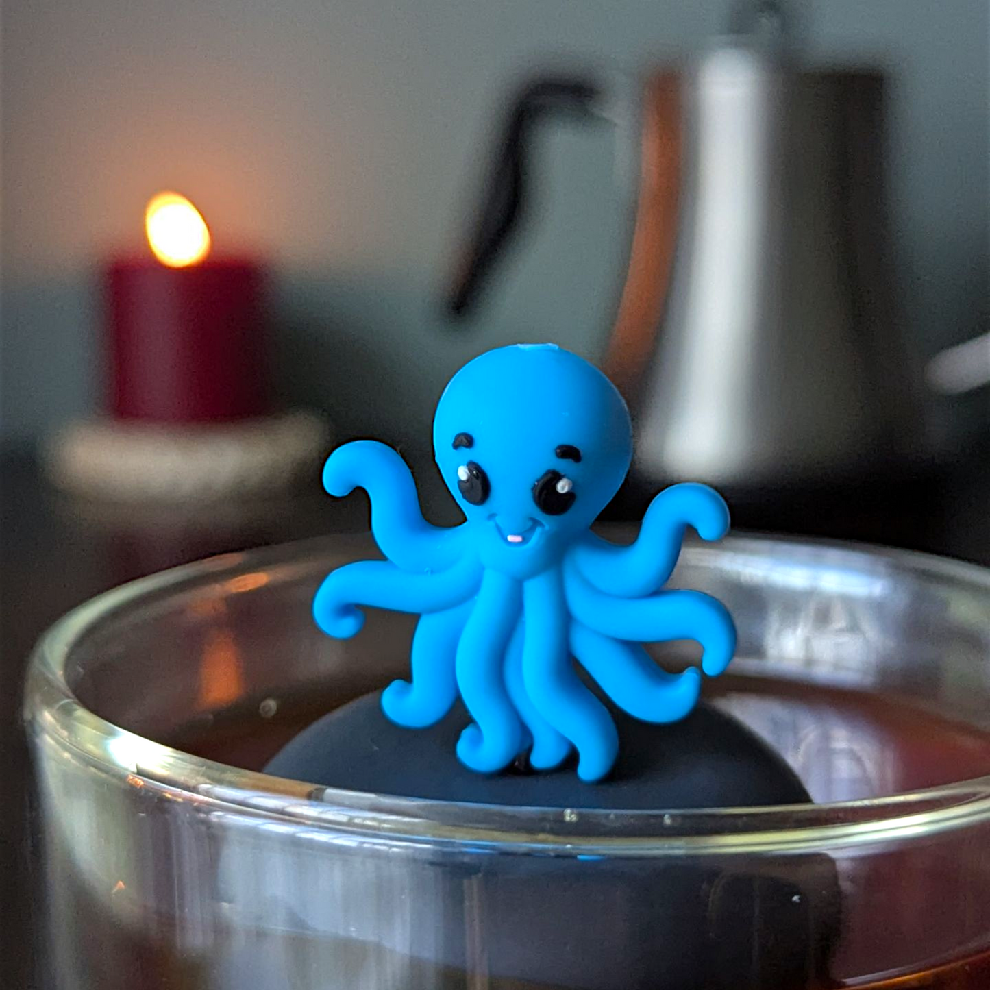 blue octopus animal tea infuser for loose leaf tea  with a fine mesh stainless steel tea infuser that reads FLOAT2INFUSE on a black base floating in a glass tea mug with a tea kettle and a candle in the background
