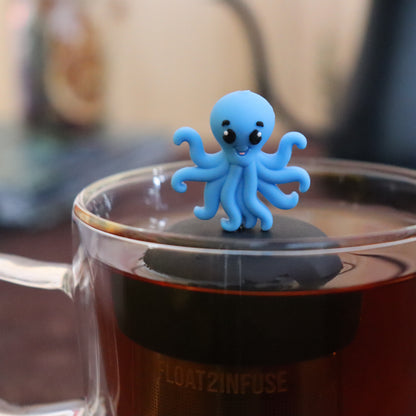 blue octopus animal tea infuser for loose leaf tea with a fine mesh stainless steel tea infuser that reads FLOAT2INFUSE on a black base  floating in a glass tea mug with tea kettle in the background