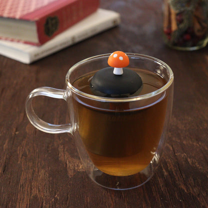 orange mushroom tea infuser for loose leaf tea  with a fine mesh stainless steel tea infuser that reads FLOAT2INFUSE on a black base floating in a glass tea mug with books and loose leaf tea in the background