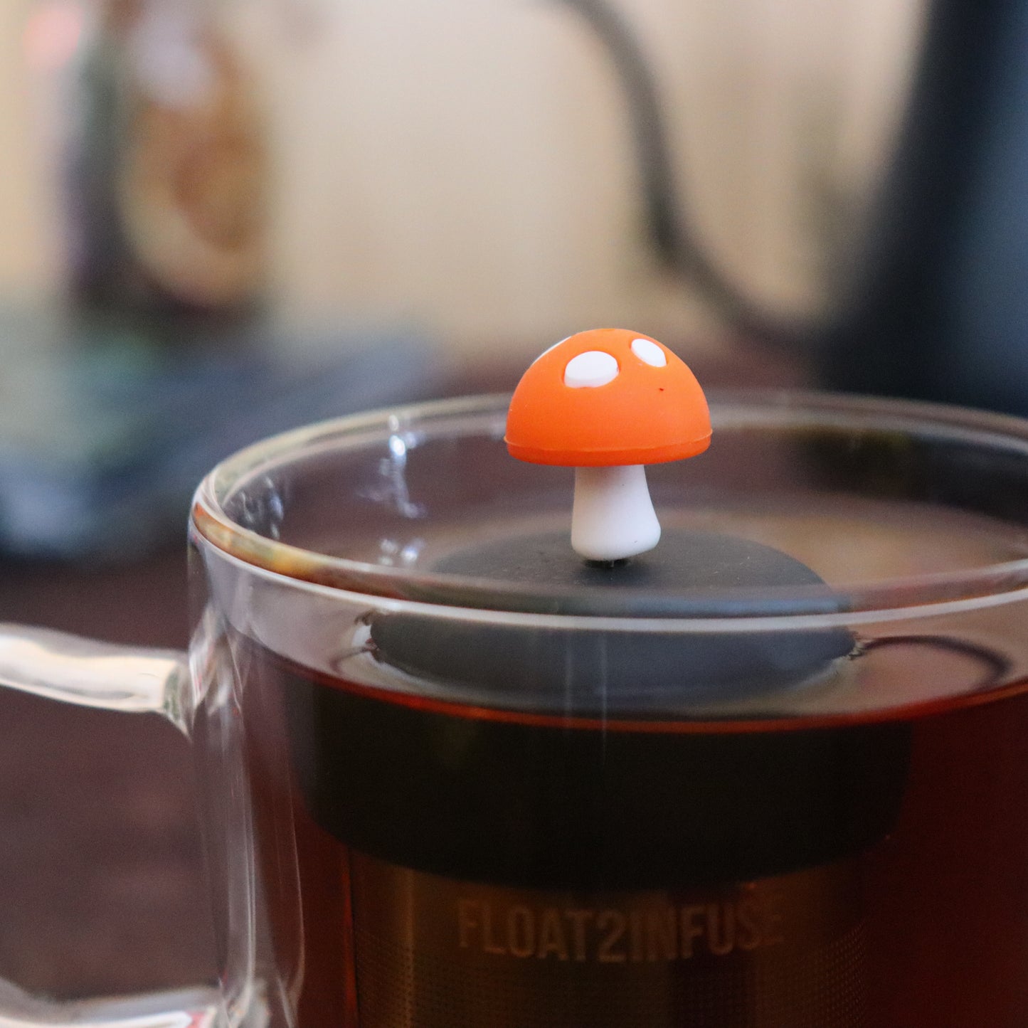 orange mushroom tea infuser for loose leaf tea with a fine mesh stainless steel strainer that reads FLOAT2INFUSE on a black base floating in a glass tea mug with tea kettle in the background