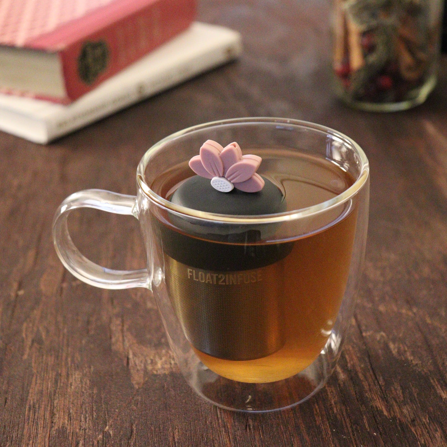 pink lotus flower tea infuser for loose leaf tea  with a fine mesh stainless steel tea infuser that reads FLOAT2INFUSE on a black base floating in a glass tea mug with books and loose leaf tea in the background