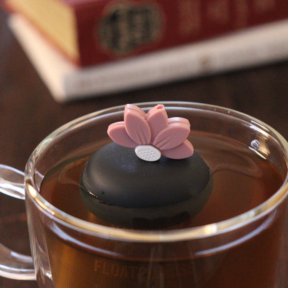 pink lotus flower tea infuser for loose leaf tea  with a fine mesh stainless steel strainer that reads FLOAT2INFUSE on a black base floating in a glass tea mug with books in the background