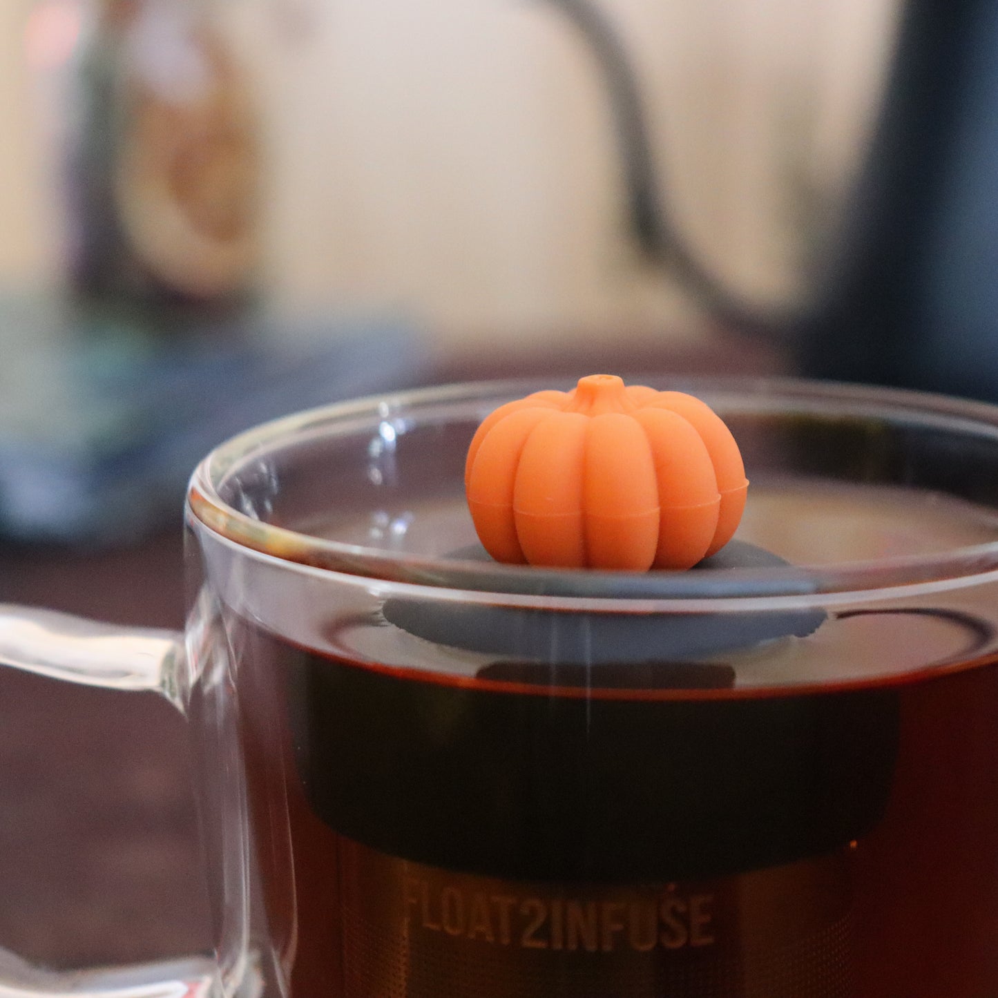 orange pumpkin tea infuser for loose leaf tea  with a fine mesh stainless steel strainer that reads FLOAT2INFUSE on a black base floating in a glass tea mug with tea kettle in the background