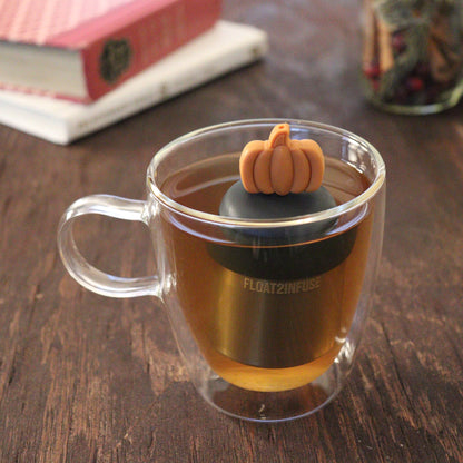 orange pumpkin tea infuser for loose leaf tea  with a fine mesh stainless steel tea infuser that reads FLOAT2INFUSE on a black base floating in a glass tea mug with books and loose leaf tea in the background