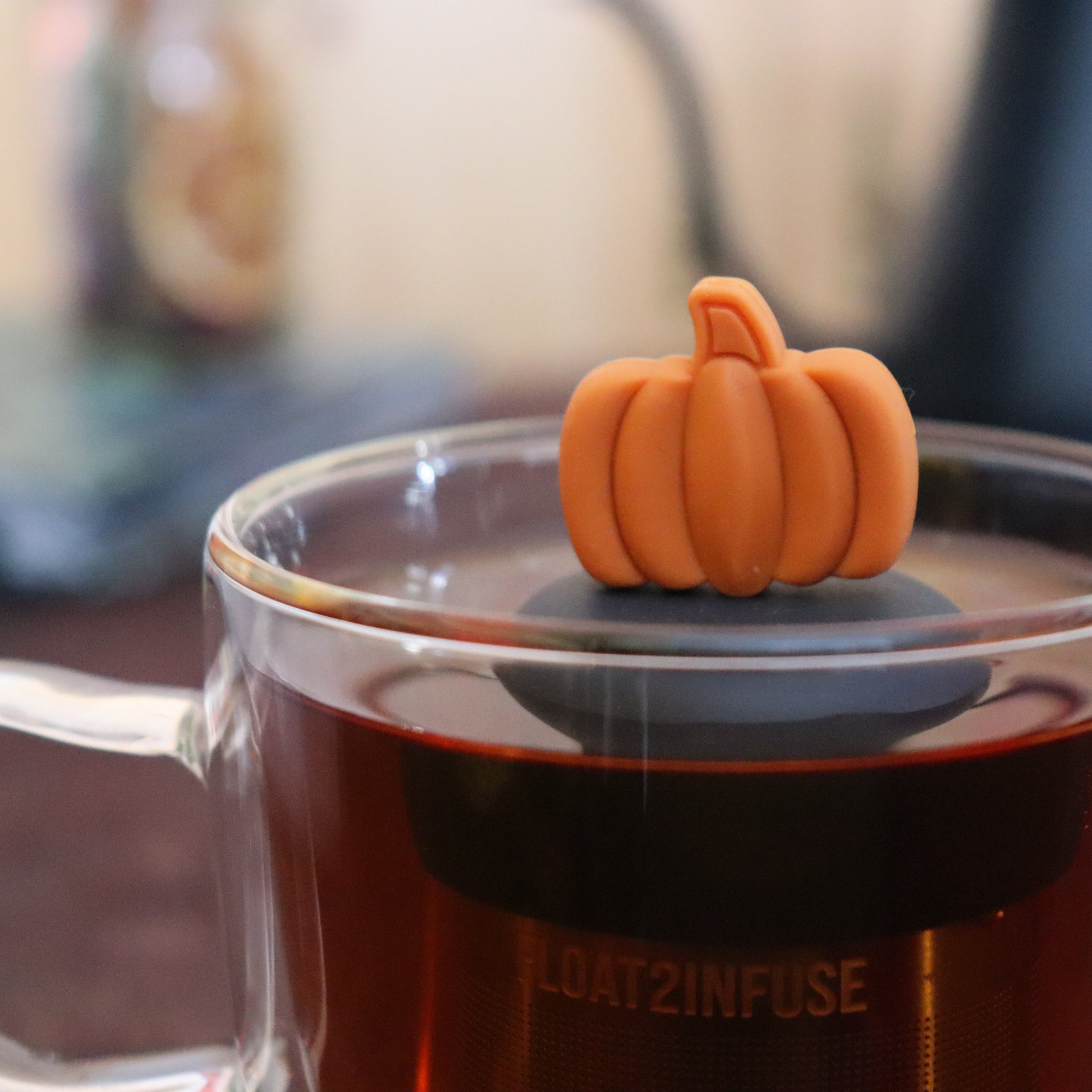 orange pumpkin tea infuser for loose leaf tea  with a fine mesh stainless steel strainer that reads FLOAT2INFUSE on a black base floating in a glass tea mug with tea kettle in the background