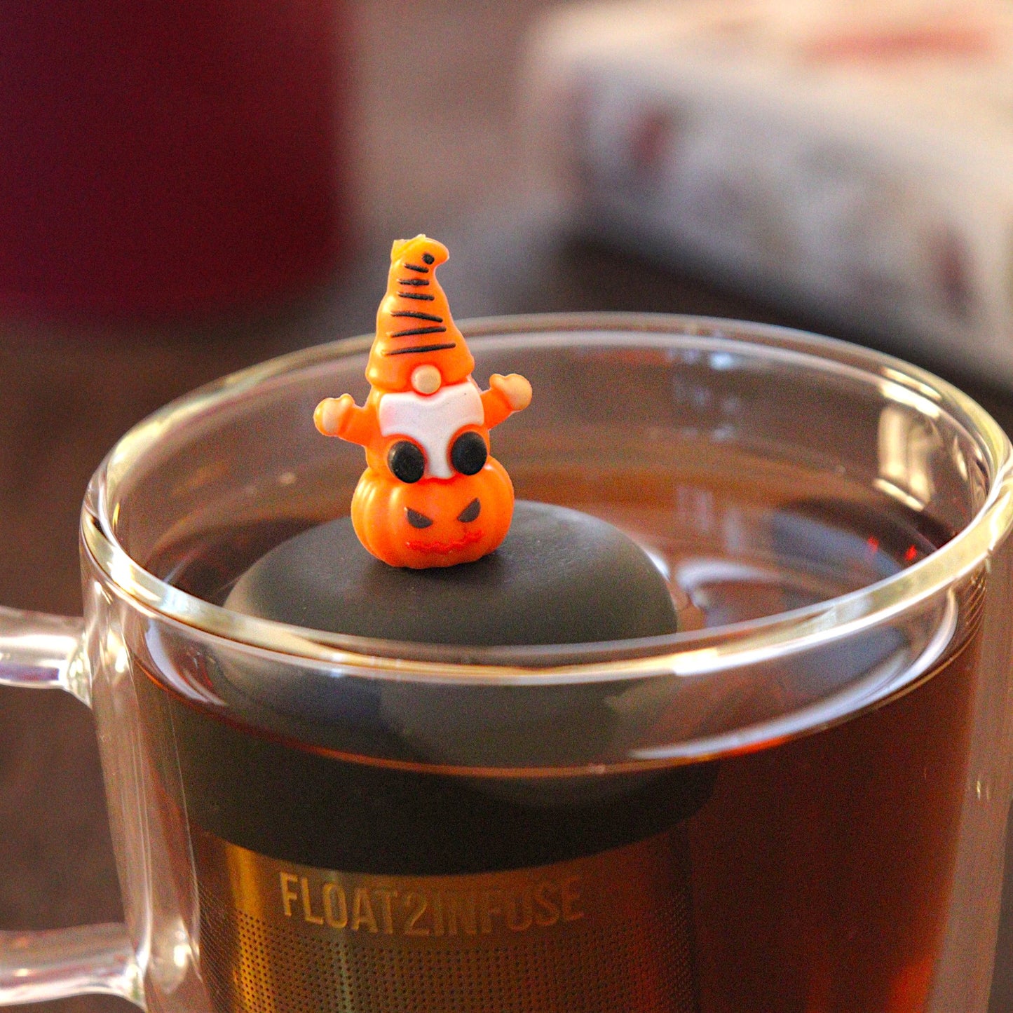 spooky orange gnome on a pumpkin tea infuser for loose leaf tea  with a fine mesh stainless steel tea infuser that reads FLOAT2INFUSE on a black base floating in a glass tea mug with a black cauldron mug and books in the background