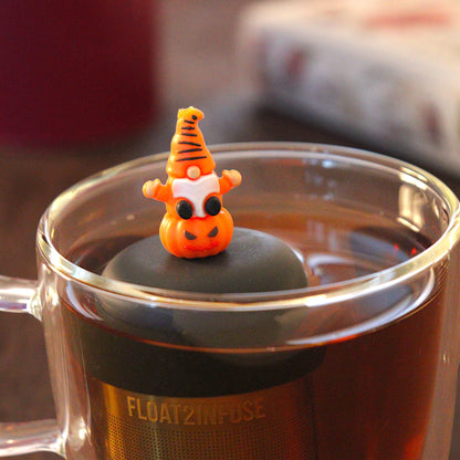 spooky orange gnome on a pumpkin tea infuser for loose leaf tea  with a fine mesh stainless steel tea infuser that reads FLOAT2INFUSE on a black base floating in a glass tea mug with a black cauldron mug and books in the background