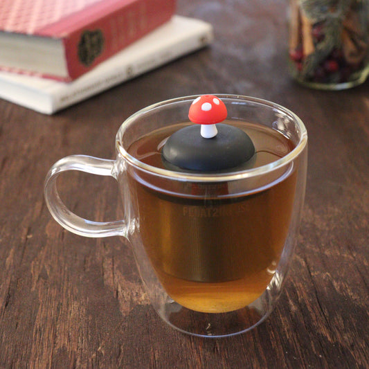 red mushroom tea infuser for loose leaf tea  with a fine mesh stainless steel tea infuser that reads FLOAT2INFUSE on a black base floating in a glass tea mug with books and loose leaf tea in the background