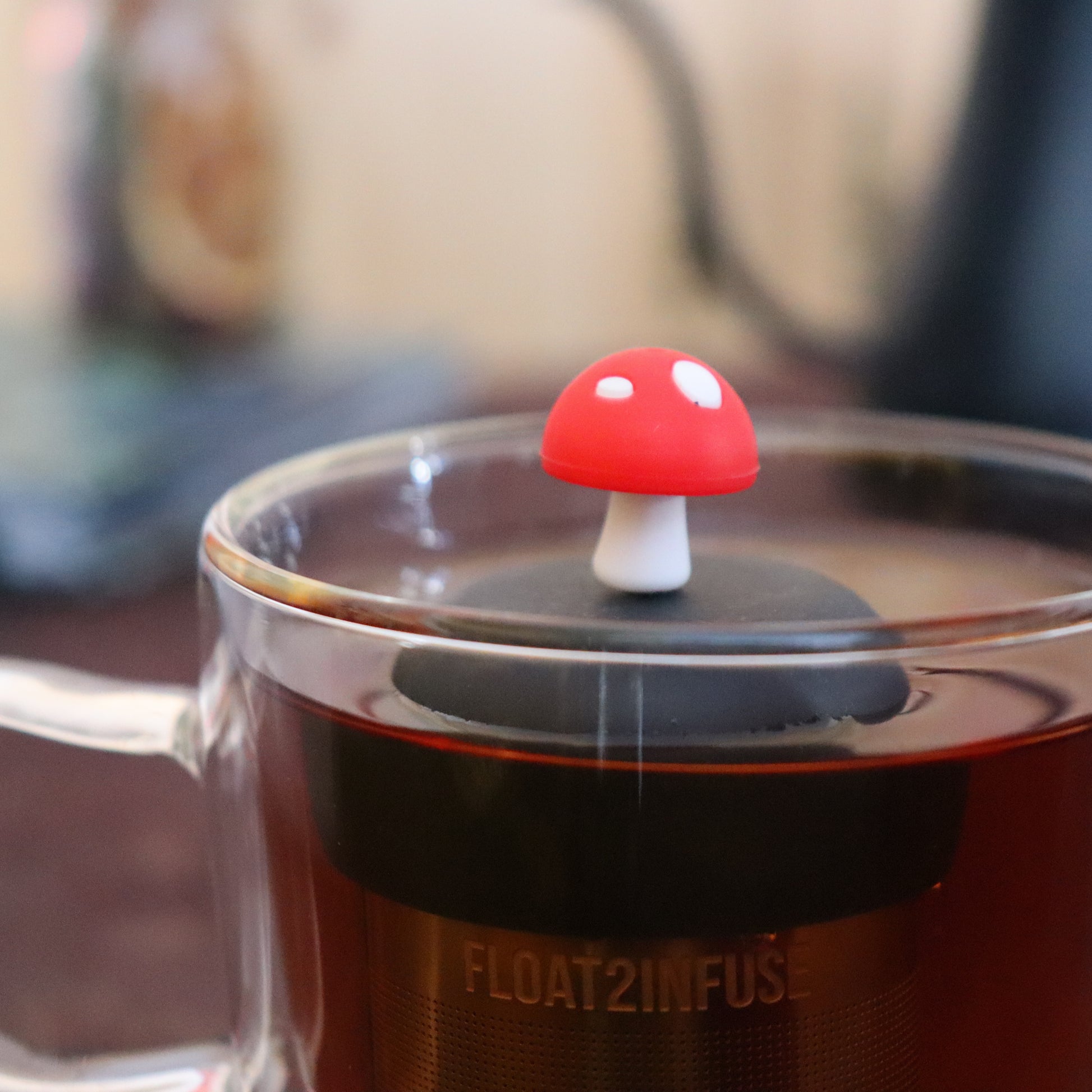 red mushroom tea infuser for loose leaf tea  with a fine mesh stainless steel strainer that reads FLOAT2INFUSE on a black base floating in a glass tea mug with tea kettle in the background