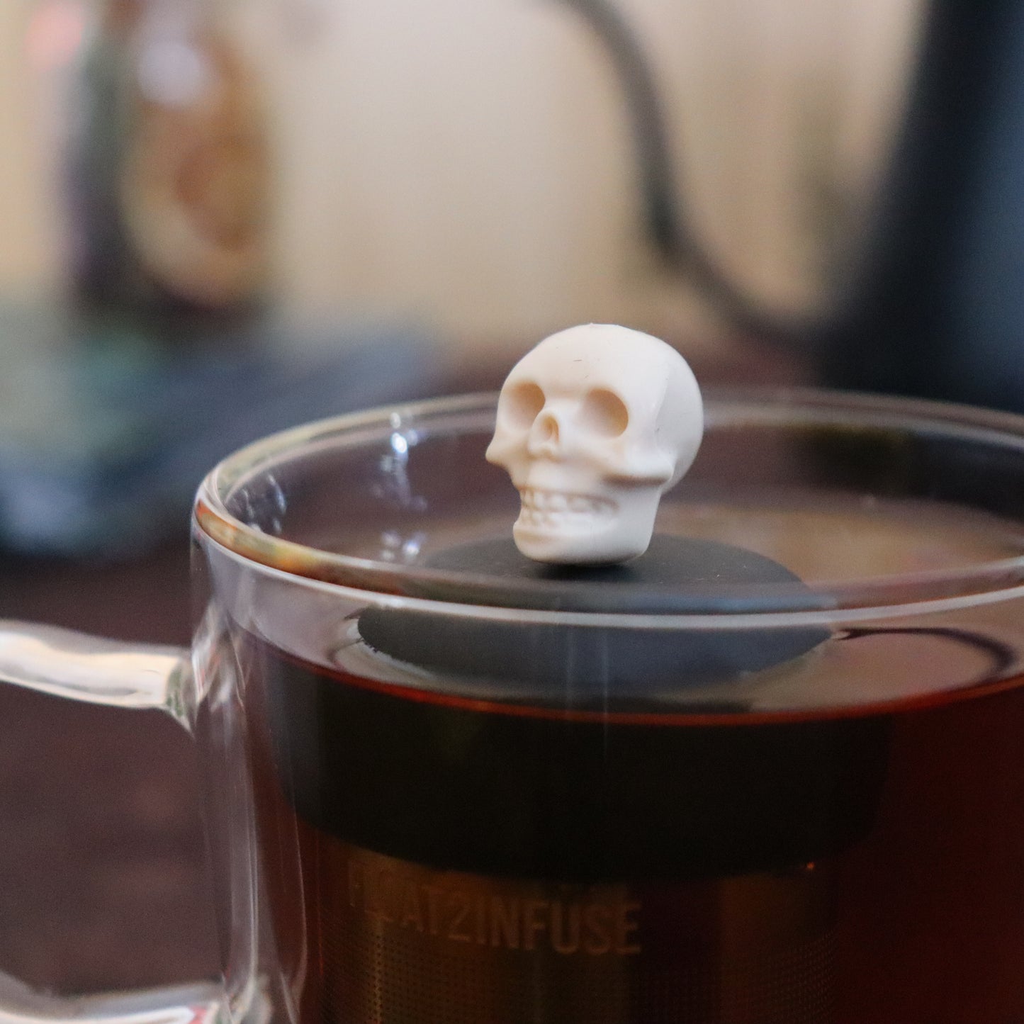 ivory skull tea infuser for loose leaf tea  with a fine mesh stainless steel strainer that reads FLOAT2INFUSE  on a black base floating in a glass tea mug with tea kettle in the background